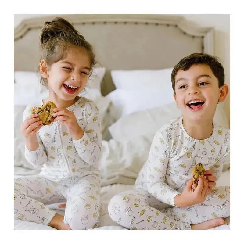 Two children wearing Bellabu Bear milk and cookies bamboo pajamas, smiling and holding chocolate chip cookies while seated on a bed.