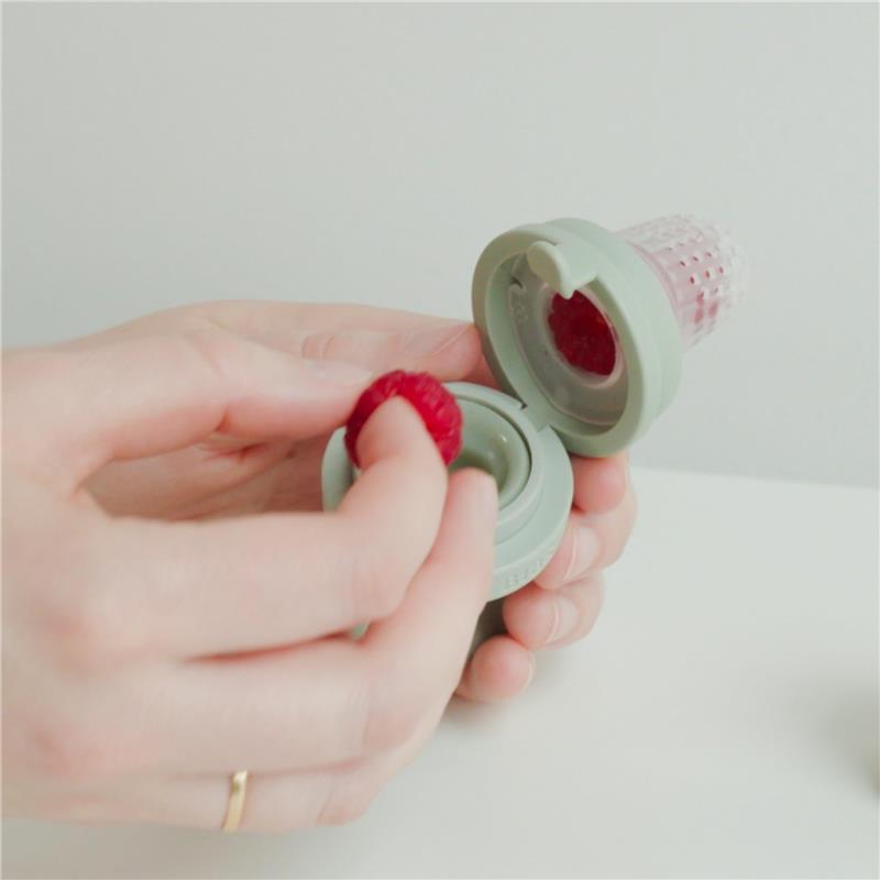 Baby Feeder in blush color held by a hand, with a raspberry being placed inside the open feeding chamber.