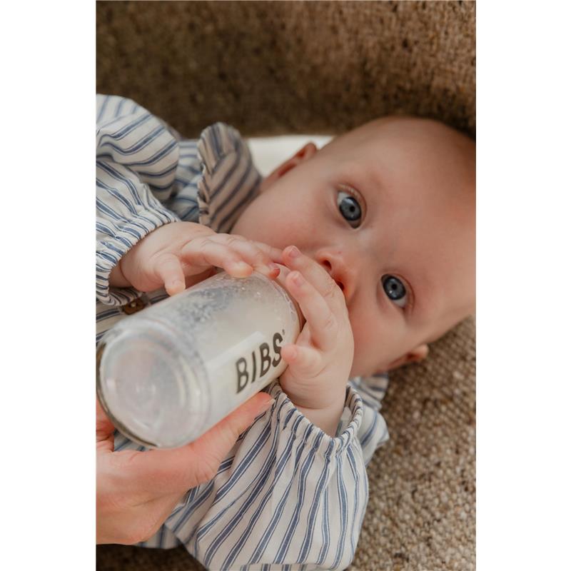 Baby holding a 150ml Bibs plastic bottle with a latex nipple, wearing a striped shirt, lying on a textured surface.