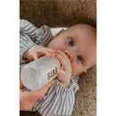Baby holding a Bibs ivory baby bottle with a latex nipple, drinking, wearing a striped outfit, and looking up.