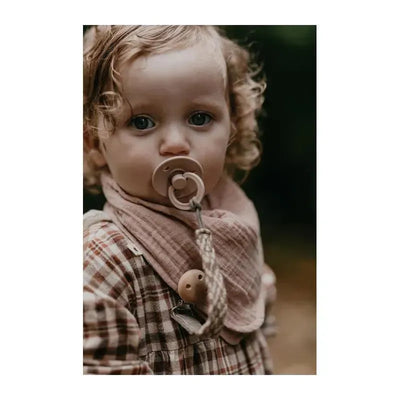 Blush bandana bib worn by a child with curly hair, holding a pacifier, set against a nature background.