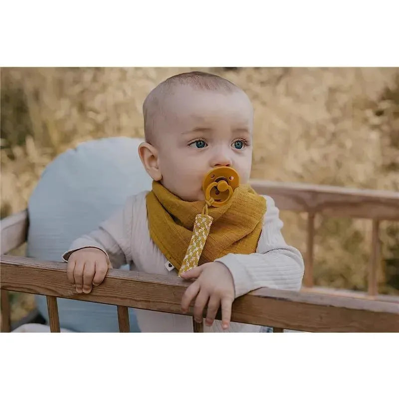 Baby with blue eyes wearing a yellow scarf holds a pacifier clipped to a dark oak and vanilla pacifier clip.
