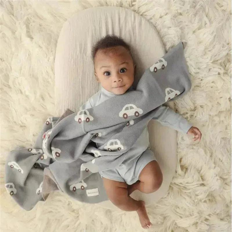 Luxury cotton baby blanket in gray with car patterns, wrapped around a smiling baby on a soft surface.