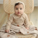 Infant sitting on a woven chair, wrapped in a mushroom-patterned luxury cotton swaddle blanket by Bleu La La.