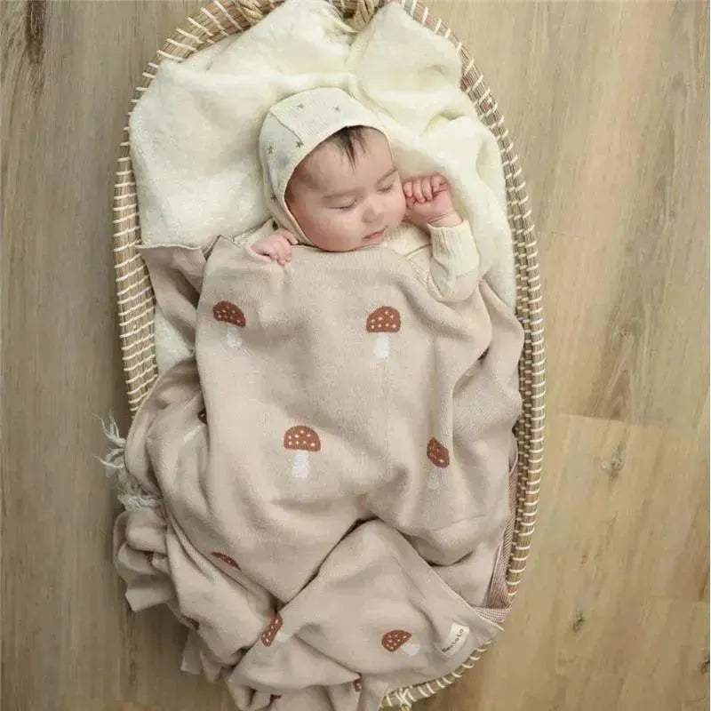 Luxury cotton swaddle blanket in mushroom camel pattern, with a sleeping baby swaddled inside, in a cozy bassinet.