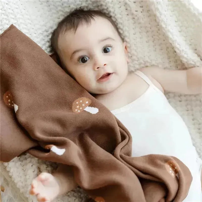 Baby boy wearing a white tank top, resting with a mushroom-patterned brown swaddle blanket on a cozy, knitted blanket.