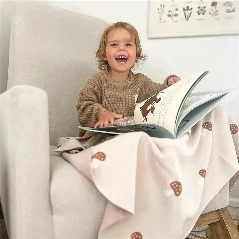 Toddler enjoying a large book while seated on a soft chair, wrapped in a pink cotton swaddle blanket featuring mushrooms.