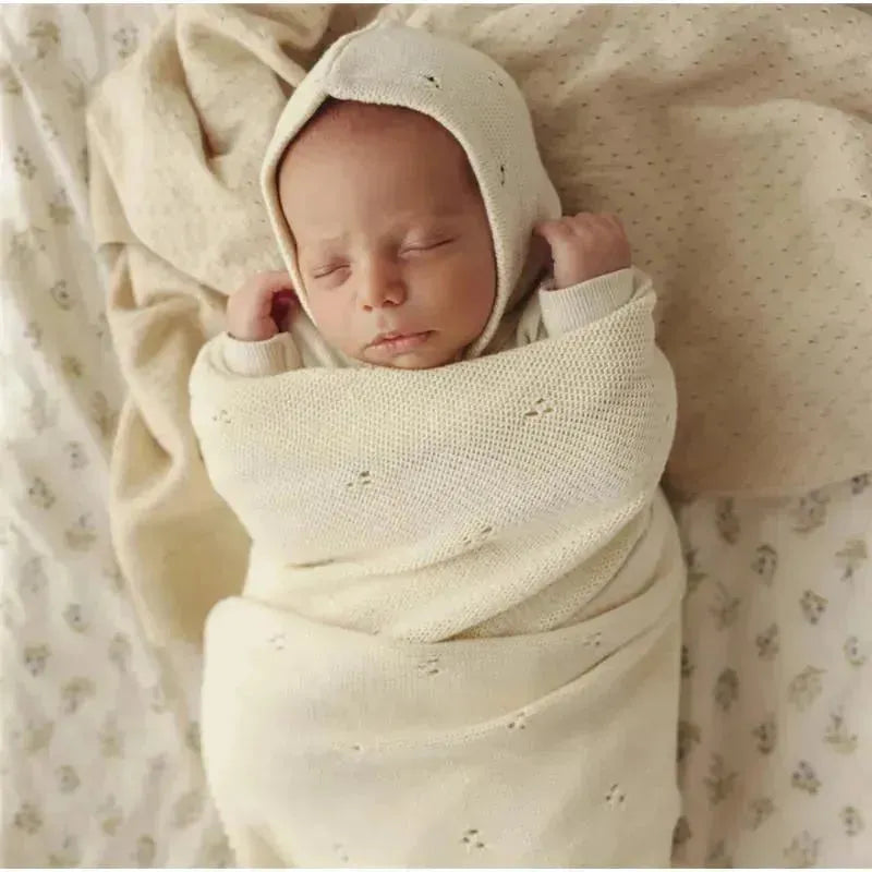 Newborn wrapped in a soft, cream-colored organic cotton blanket with a matching bonnet, resting peacefully on a patterned background.