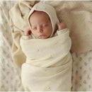 Newborn wrapped in a soft, cream-colored organic cotton blanket with a matching bonnet, resting peacefully on a patterned background.