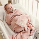 A baby swaddled in a pink pointelle blanket from Bleu La La, resting in a crib with a pacifier and a plush toy.