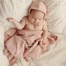 Baby wrapped in a pink organic cotton pointelle swaddle blanket, wearing a matching bonnet, resting on a soft surface.
