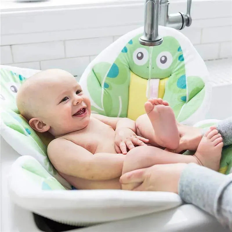 A happy baby sits in a frog-themed Blooming Baby bath seat, with water gently flowing from the faucet above.