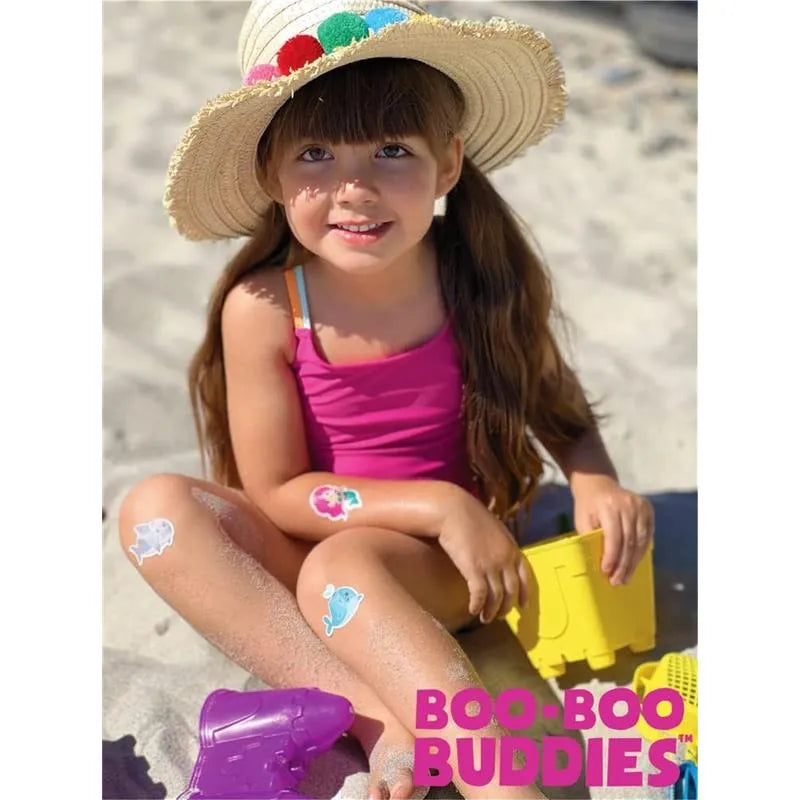 Young girl in a pink swimsuit with a sun hat sitting in sand, displaying colorful adhesive bandages on her legs.