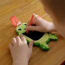 Child drawing on a green turtle-shaped doodle board with a red stylus on a wooden surface.