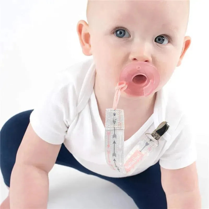 Baby with blue eyes wearing a white shirt uses a pink pacifier attached to a pink arrows-patterned clip.