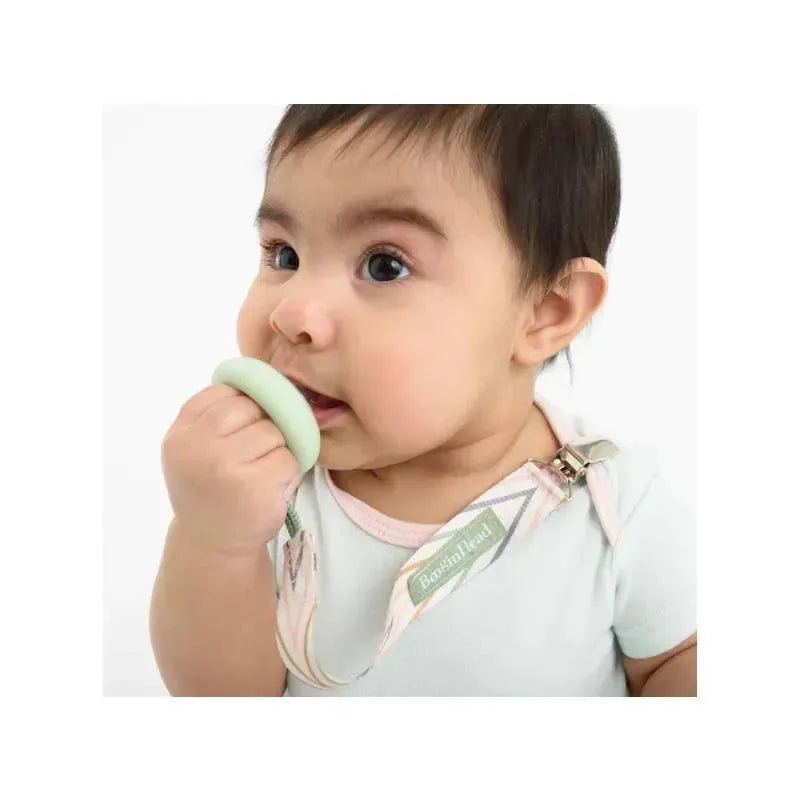 Baby using a Booginhead pacifier clip in geometric pinks while holding a green silicone pacifier, wearing a light shirt.