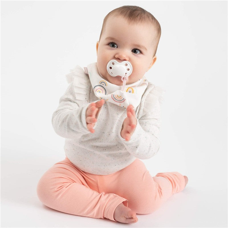 Baby wearing a pink bandana bib with rainbow print and holding a pacifier, sitting and clapping hands.