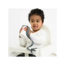 A young child in a high chair holds a white silicone sippy cup with a charcoal gray strap attached.