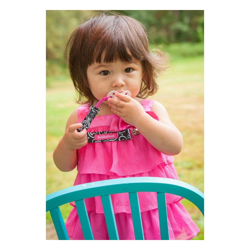 Toddler in a pink dress holding a Booginhead Sippie Grip with a black flourish design, seated by a teal chair.