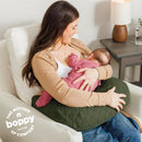 A mother uses a Boppy Nursing Pillow Luxe in moss quilted fabric while breastfeeding her baby, both relaxed on a couch.