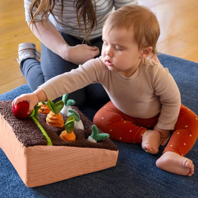 Toddler playing with the Boppy Learn and Grow Garden, engaging with plush vegetables and soil on a blue rug.