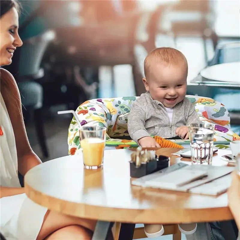A happy baby sits in a colorful Boppy shopping cart cover, engaged with a toy, while a woman smiles nearby at a café table.
