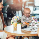 A happy baby sits in a colorful Boppy shopping cart cover, engaged with a toy, while a woman smiles nearby at a café table.