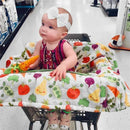 Infant in a shopping cart covered with a colorful Boppy cart cover featuring fruit and vegetable designs, holding a toy carrot.