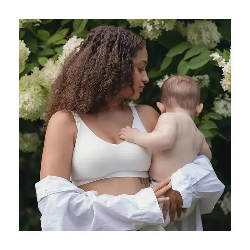 A woman with curly hair holds a baby while wearing a white Bravado Body Silk Seamless Nursing Bra against a floral background.