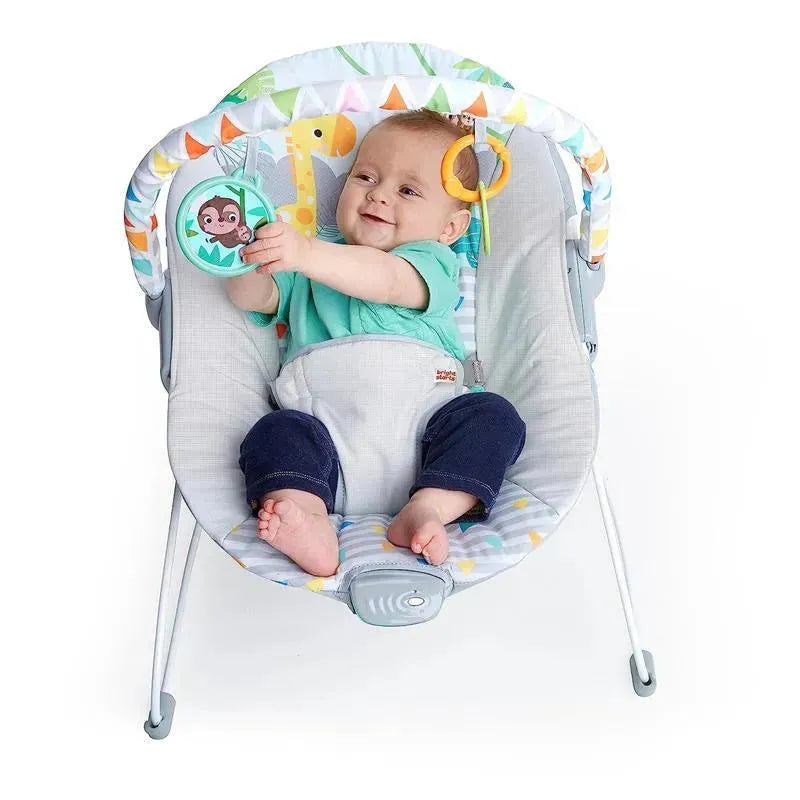 Baby in a Bright Starts bouncer seat reaches for a hanging toy, smiling, with a colorful, soft fabric seat design.
