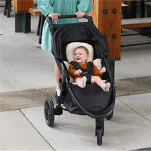 A parent pushes a black stroller with a smiling baby wearing an orange outfit and white socks, on a paved walkway.