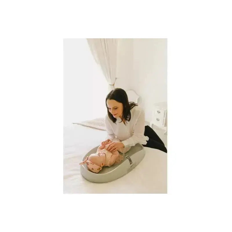 Woman assisting a baby on a Bumbo changing pad in cool grey on a white bed, demonstrating safe diaper changing.