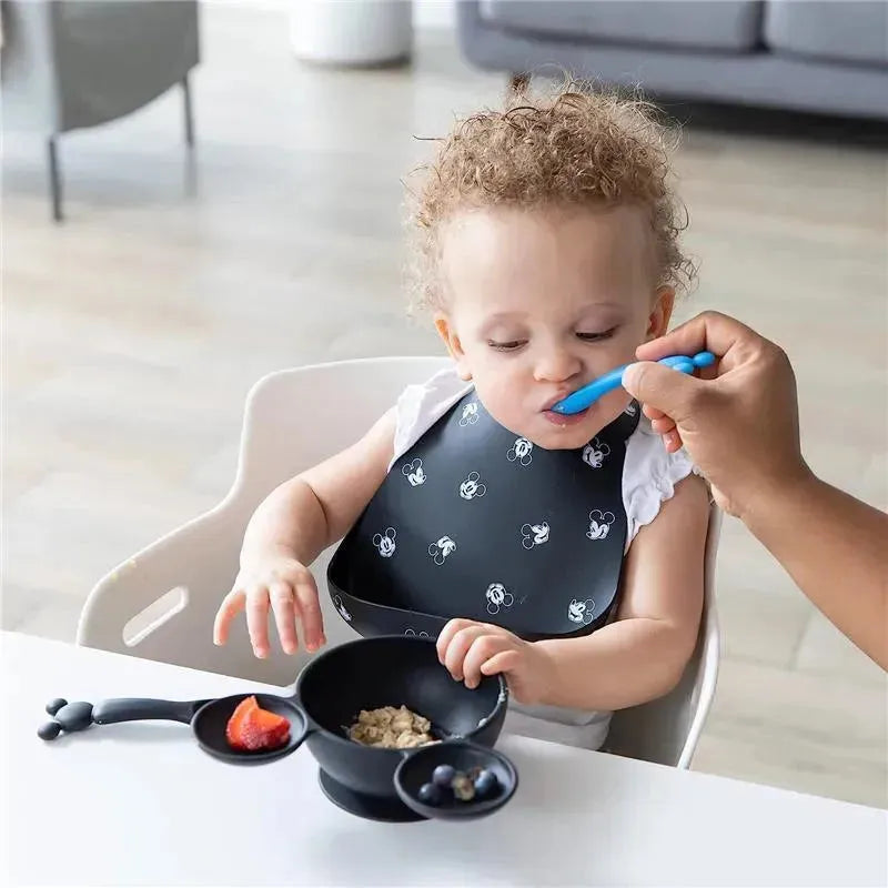 A toddler with curly hair wearing a Mickey Mouse bib is being fed with a blue spoon. A bowl contains oatmeal and fruit.
