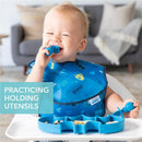 A child wearing a Batman-themed bib smiles while practicing holding blue silicone utensils and sitting at a high chair.