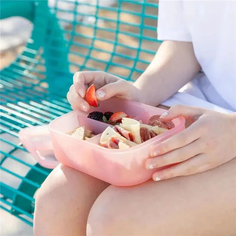 Bumkins pink bento box with three compartments filled with snacks, held by a child sitting on a bench.