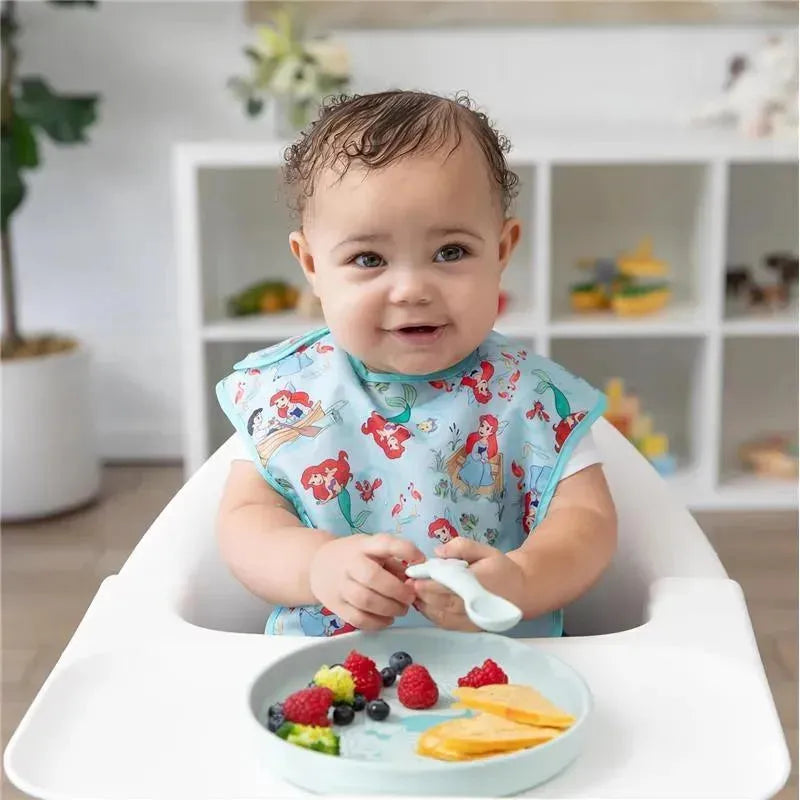 Baby wearing a blue Ariel-themed bib, holding a spoon, with a plate of fruit including berries and orange slices.