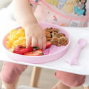 Silicone plate and spoon set featuring a pink design, with a child picking food, including strawberries and scrambled eggs.