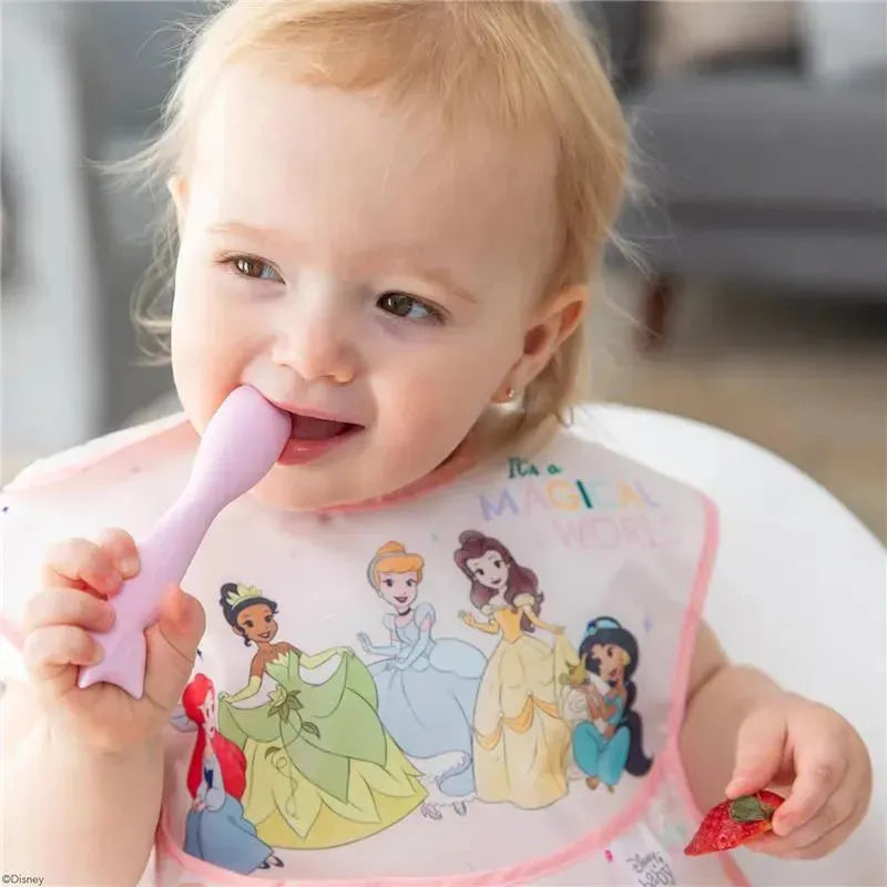 Toddler enjoying a pink spoon while wearing a Disney princess bib, with a strawberry in her other hand.