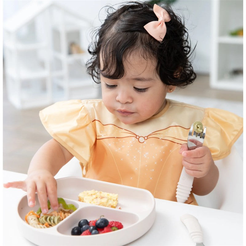 Child wearing a Belle-themed ruffle apron bib reaches for food on a divided plate with pasta, berries, and cake.