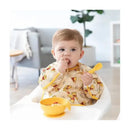 Baby in a yellow Winnie the Pooh bib, holding food and a spoon, sitting at a high chair with a yellow bowl of food.