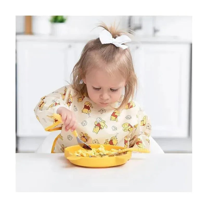 Toddler wearing a Bumkins Disney Winnie the Pooh sleeved bib, focused on eating from a yellow plate.