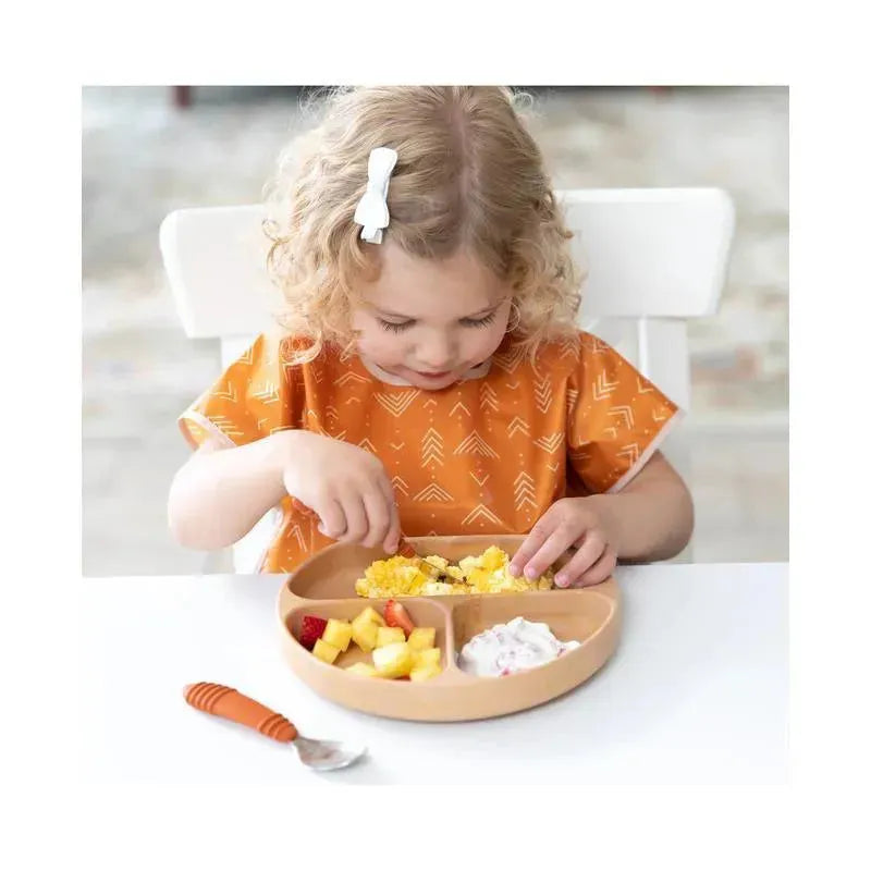 A young child in an orange patterned bib engages with a divided beige plate filled with food, including fruit and yogurt.