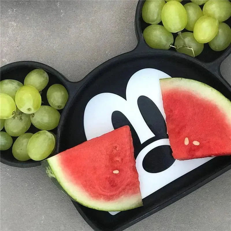 Mickey Mouse silicone grip dish with green grapes and watermelon slices arranged inside.