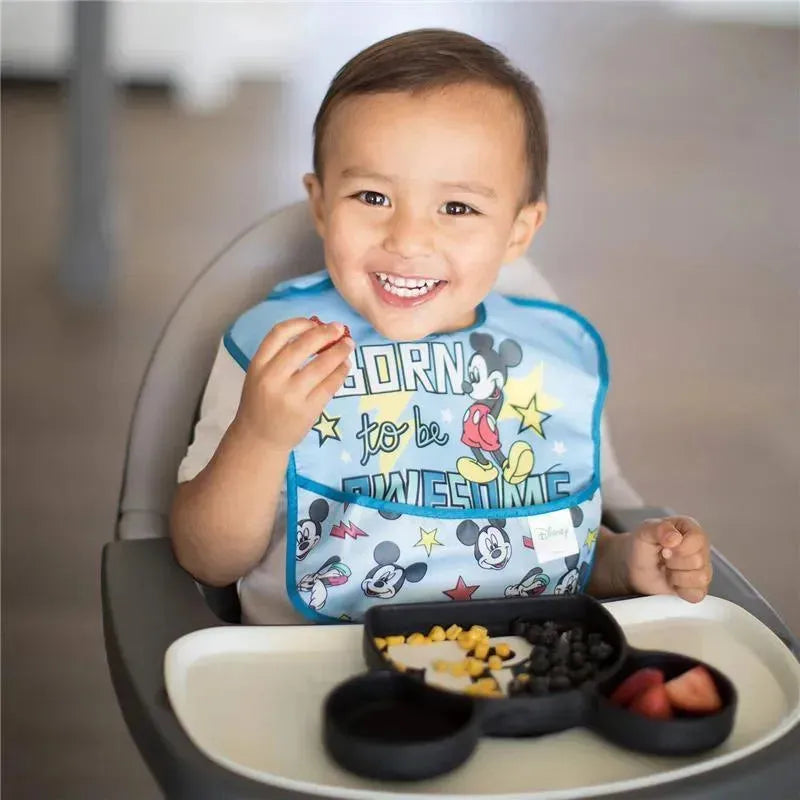 A smiling child in a high chair wears a Mickey Mouse bib and enjoys food from a black silicone grip dish.
