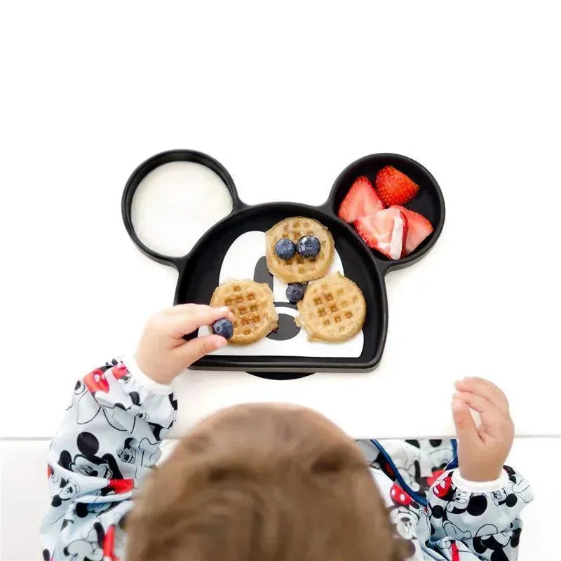 Mickey Mouse-shaped silicone grip dish with waffles, blueberries, and strawberries, held by a child in a Mickey-themed outfit.