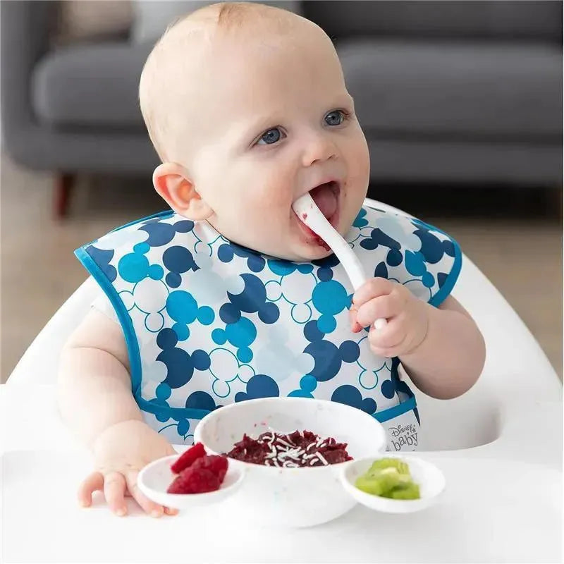 Baby wearing a Mickey Mouse bib, using a spoon to eat from a bowl with red food and surrounded by fruit in small dishes.