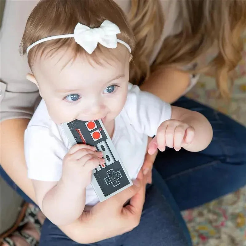Baby with blue eyes chewing on a grey and black Nintendo controller silicone teether while being held.