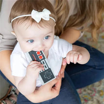 Baby with blue eyes chewing on a grey and black Nintendo controller silicone teether while being held.