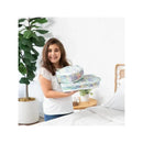 A girl smiling while holding two Bumkins packing cubes in pastel Aloha design, in a bright room with plants and bedding.