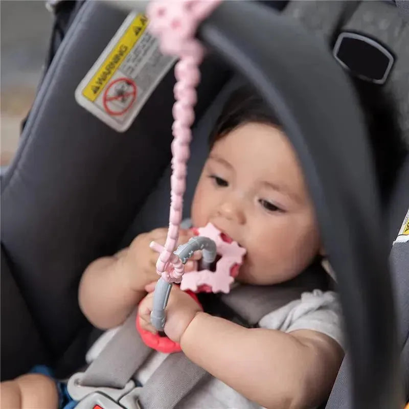 Baby in a car seat holding a pink silicone teether attached to a pink tether, designed to keep toys within reach.